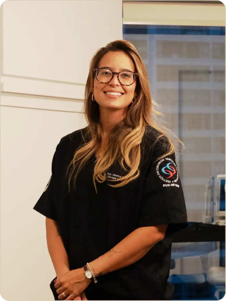 Retrato de Jessica Santos, profissional da área da saúde, sorrindo para a câmera em um ambiente clínico. Ela usa óculos, cabelos longos com mechas claras e veste jaleco preto com identificação de cirurgiã vascular bordada no peito e na manga. Está em pé, com as mãos à frente do corpo, em um consultório bem iluminado, com equipamentos médicos ao fundo e uma janela ao lado.