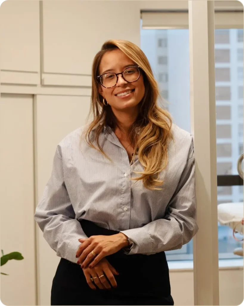 Retrato de Jessica Santos sorrindo para a câmera em um consultório. Ela usa óculos, camisa social listrada em tons claros e calça preta, com os cabelos longos soltos e levemente ondulados. Está apoiada em uma divisória, com as mãos cruzadas à frente do corpo. Ao fundo, há uma janela com vista para edifícios e elementos do ambiente clínico, em um espaço bem iluminado e acolhedor.