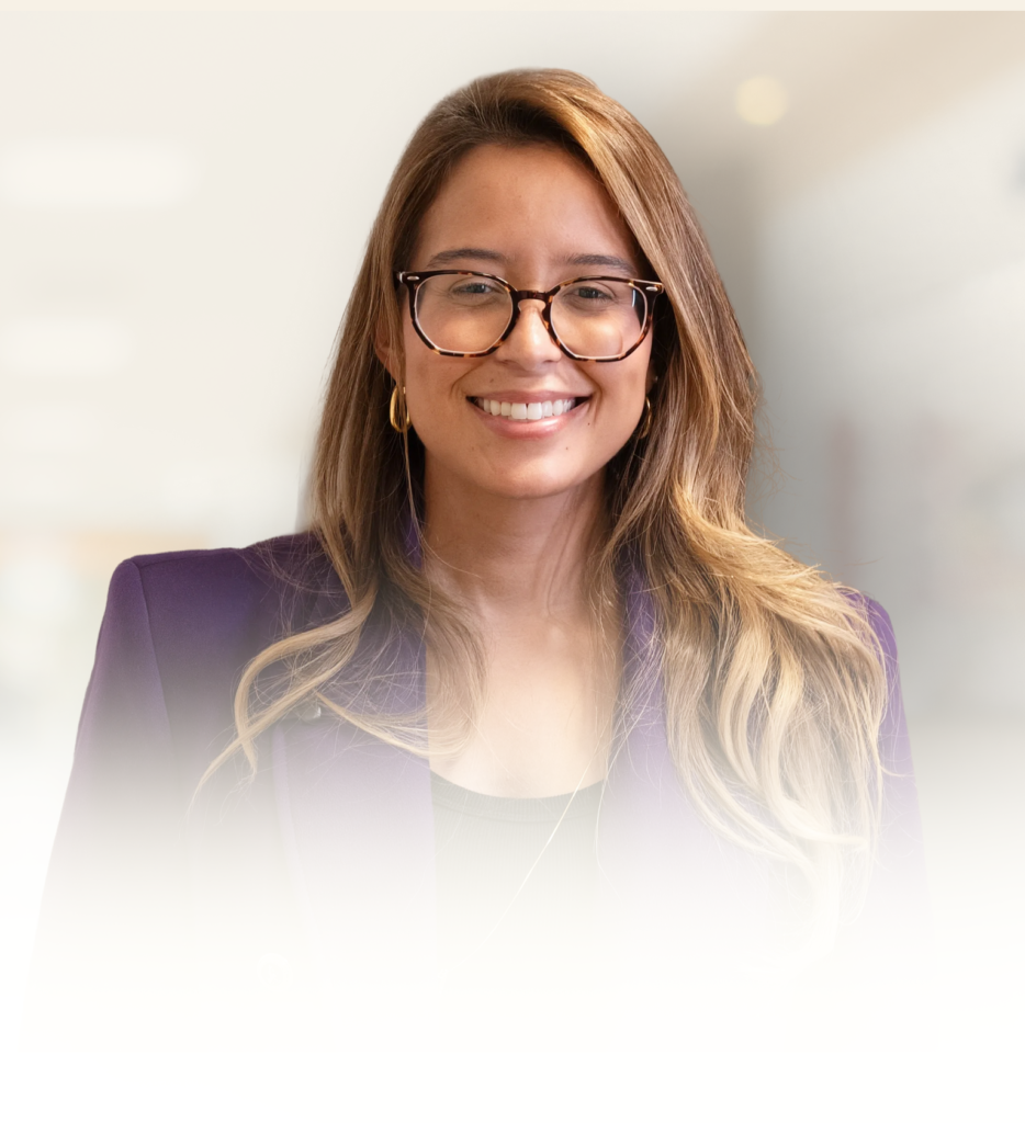 **Texto alternativo (alt text):** > Retrato de Jessica Santos sorrindo para a câmera. Ela usa óculos de armação escura, brincos de argola e blazer roxo, com os cabelos longos e lisos em tons castanhos com mechas claras. O fundo desfocado em tons claros cria um efeito suave e profissional, destacando seu rosto e transmitindo uma imagem acolhedora e confiante.